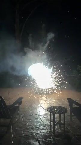 A close-up shot captures fireworks exploding on a patio at night. Sparks fly outwards from a central bright light, illuminating the wet, stone patio. In the foreground, two empty adirondack chairs and a small table are silhouetted against the light. The surrounding trees and sky are dark, indicating nighttime.