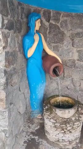 The video shows a water feature comprised of statues and a stone basin. A statue of a woman in a blue dress and headscarf is pouring water from a brown clay jar into a stone basin. Next to the basin is a statue of a man with long brown hair and a beard, wearing a brown robe, and sitting on a rock. A single-use face mask is on the ground near the statues. The background is a stone wall with a blue ceiling. The water feature is likely a religious display.