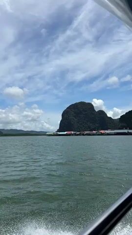 The video shows a body of water with a boat in the foreground, creating a wake. In the background, there is a lush green, mountainous island with a village of colorful houses built on stilts over the water. The sky is blue with scattered white clouds. The scene appears to be a tropical location.