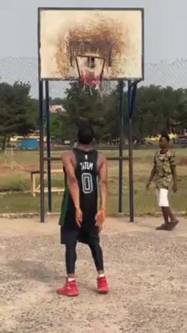 A medium shot depicts a man wearing a basketball jersey with the name "Tatum" and number "0" on the back, standing on a basketball court. He is facing a basketball hoop and about to shoot a basketball. The basketball hoop is weathered with rust and peeling paint. Another man stands off to the side in the background, wearing a patterned shirt and white shorts, watching the first man. The basketball court appears to be outdoors, with green grass and trees visible in the background. A section of chain-link fence runs along the back of the court. There is a bench in the background near the fence. The scene is well-lit, suggesting it's daytime. The camera captures the moment just before the man releases the ball towards the hoop.