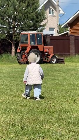 A toddler wearing a light gray vest, blue pants, and a beige beanie stands on a green lawn.  In the background, there's an old orange tractor parked near a brown fence. Beyond the fence, one can see a two-story house with a white roof and a log cabin style building. A tall pine tree stands to the left of the tractor and the sky is blue with occasional clouds. A utility pole with wires is also visible. The toddler is facing away from the camera, observing the tractor.