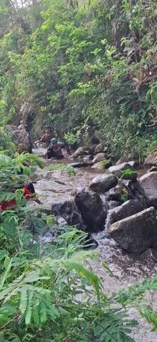 The video shows a group of people in a shallow river. The riverbed is rocky, and the water is clear and moving. Lush green vegetation lines the banks of the river. Some individuals are partially submerged in the water, appearing to be washing themselves or fetching water. Others are seated or standing on the rocks along the riverbank. The atmosphere seems relaxed and everyday.