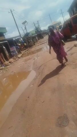 A woman wearing a purple and pink patterned dress walks down a muddy, uneven road. The road is filled with puddles, and there are several buildings and vehicles in the background. The sun is shining, casting a long shadow from the woman.