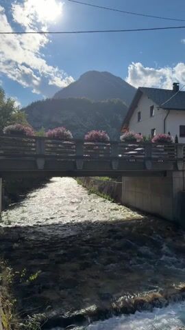 The video shows a wide shot of a fast-flowing river passing under a bridge. The bridge is decorated with planters filled with pink flowers. In the background, a mountain rises under a bright, sunny sky with some clouds. A house with a white facade and a dark roof is visible to the right of the mountain. The sun is shining brightly, creating dappled light on the water and casting shadows on the bridge. The overall scene is scenic and peaceful, with a focus on nature and a village setting. There are no people or animals visible in the video. The camera appears to be stationary.