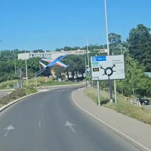 The video shows a static display of a blue, white, and red jet airplane affixed on a vertical post, situated within a roundabout intersection. The camera, seemingly mounted within a vehicle, approaches the roundabout along a two-lane asphalt road. The day is sunny with a clear blue sky. Road signage indicates directions and destinations.  Vegetation and buildings are visible in the background. No people are discernable. The vehicle continues driving without stopping or turning within the frame.