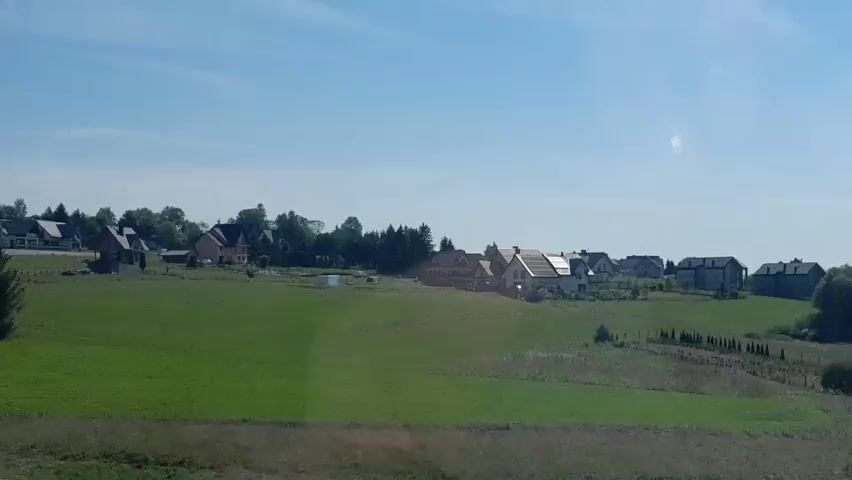 The video shows a rural landscape with several houses visible on a gentle slope. A large, green field dominates the foreground. The sky is clear and blue with a few wispy clouds. The camera appears to be moving, possibly from a vehicle, as indicated by a blurred railing in the lower left corner of the first frame. The houses are scattered across the hilltop, and some appear to have solar panels on their roofs. There are trees and some smaller structures interspersed with the houses. The overall impression is one of a tranquil countryside setting on a sunny day.