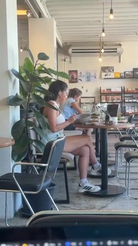 The media file shows an indoor scene, likely a cafe or restaurant in Choeng Mon, Thailand. Two women are seated at a table, and one of them is eating. A large potted plant with dark green leaves is in the foreground, obscuring part of the scene. The woman in the foreground is wearing a white tank top and light blue shorts, with her legs crossed. She is looking down at her phone. The woman in the background is wearing a light blue shirt and is eating from a bowl. A laptop is open on the table in front of her, and there are drinks and other items on the table.

The setting appears bright and airy, with fluorescent lighting and exposed ceiling beams. Shelves with various products are visible in the background, suggesting a retail component to the establishment. The overall mood seems relaxed and casual. The time of day is likely daytime, given the natural light filtering in. There is no indication of the weather.

No text is clearly legible in the media file, though some posters or artwork are visible on the wall in the background.