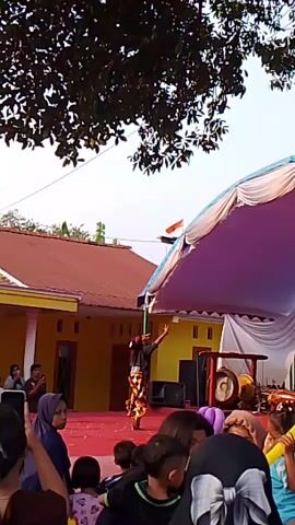 A crowd watches a performer in traditional dress on a red stage under a tent. The performer moves dynamically, facing the audience. To the right of the performer, musical instruments, possibly gongs, are visible on the stage. Behind the stage, a yellow building with a red roof is visible. Overhead, tree branches with leaves are present. The audience members are varied in age, with some individuals holding up cell phones to record the performance. The scene appears to be outdoors, possibly during a daytime event or festival.