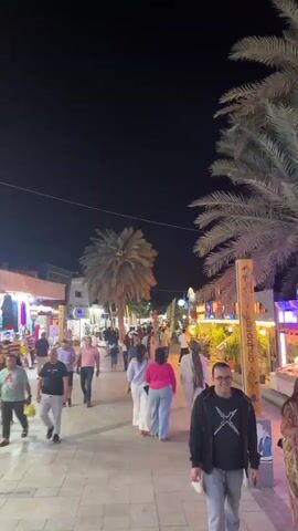 A busy street is shown at night, lit by streetlights and storefronts. People of various ages and ethnicities are walking along the street. The street is lined with shops selling clothing and souvenirs. Palm trees line the street.