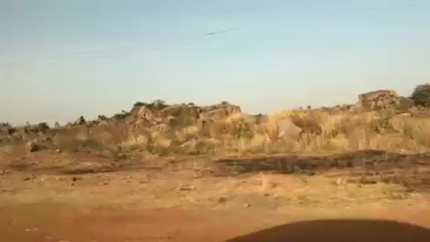 The video shows a landscape as seen from a moving vehicle. The terrain is predominantly dry and dusty with a reddish-brown hue. There are clusters of large rocks and sparse vegetation visible. An electric pole is noticeable at one point along the roadside. The sky is pale blue. The overall environment appears arid.