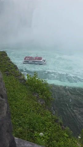 A tour boat with many passengers wearing red ponchos is moving through choppy, turquoise water near a cliff covered in grass. Mist is visible in the background, obscuring a large waterfall. The boat appears to be approaching the base of the waterfall.