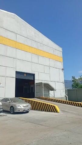 The video shows the exterior of a large, rectangular industrial building. It's primarily light gray with a horizontal yellow stripe running along its upper half. The building has a large, open loading bay with a vehicle parked nearby. There is a ramp leading up to the loading bay with yellow and black hazard stripes. Multiple vehicles are parked in front of the building. The sky is blue with scattered clouds. A sign on the building displays "Oliven". A truck is parked in front of the building. Several people are visible around the truck.