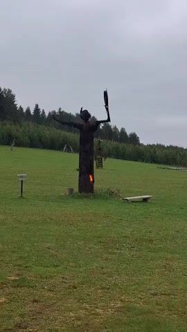 The video shows a wide shot of a field with a metal sculpture in the center. The sculpture is in the shape of a person with arms outstretched, and flames are visible within the torso area of the sculpture. The field is green and grassy, and there are some trees and bushes in the background. The sky is overcast and gray. There is a small wooden sign to the left of the sculpture, and a simple wooden bench to the right. Another sculpture is visible far in the left background. The camera is static and does not move throughout the video.