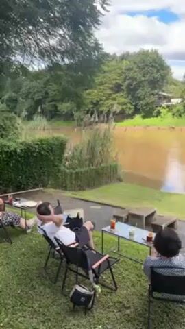 Two people are sitting in chairs on a grassy lawn, facing a river. The river is wide and muddy, with trees lining the opposite bank. There are more chairs and tables set up along the riverbank, and a small building is visible in the distance. The sky is partly cloudy. One person is leaning back in their chair with their hands behind their head, while the other is sitting upright. There is a small table with drinks on it between them.