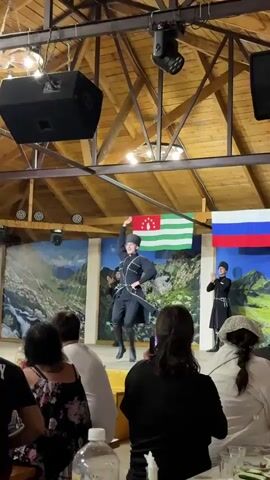 The video captures a cultural performance on a wooden stage. In the foreground, the backs of several audience members are visible, some holding up their phones to record. The stage itself has a backdrop depicting a mountainous landscape. Prominently displayed on the stage are two flags: the flag of Abkhazia and the flag of Russia. The central focus of the performance is a male dancer in traditional attire, including a dark, elaborately decorated tunic, trousers, and a distinctive cap. He is performing a dynamic dance with energetic movements and arm gestures. To his sides, two other performers, also in similar traditional attire, are clapping and performing synchronized movements. Stage lighting, including blue spotlights, illuminates the performers and the backdrop. Above the stage, large black speakers are mounted on the wooden ceiling structure. The overall environment suggests an indoor venue, possibly a restaurant or cultural center, hosting a traditional dance performance.
