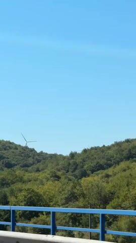 The video shows a view from a moving vehicle, looking out over a lush green forested hillside under a clear blue sky. In the distance, several wind turbines are visible, their blades appearing still against the horizon. A highway guardrail with blue top bars runs along the bottom of the frame, indicating the perspective is from a road or bridge. The road curves into the distance among the trees. The overall impression is one of a scenic drive through a rural, possibly hilly, landscape.