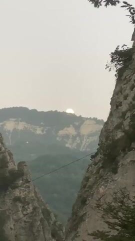 A video captures a mountainous landscape with the sun partially obscured by a mountain peak. The sun appears as a bright, glowing orb near the top of the distant mountain. The foreground features two large, rocky cliffs on either side, creating a natural frame for the scene. Lush green foliage covers the slopes of the mountains in the mid-ground. A thin, dark cable stretches horizontally across the frame between the two prominent cliffs in the foreground, suggesting a man-made element within the natural environment, possibly a zipline or power line. The sky is overcast with a light, hazy glow, indicating either dawn, dusk, or a heavily clouded day. The overall atmosphere is serene and majestic, highlighting the grandeur of the natural terrain.