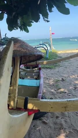 The video shows a close-up of a traditional Indonesian jukung boat on a sandy beach. The boat is made of wood and has outriggers. A woven conical hat is placed on top of the boat. In the background, the ocean and a clear blue sky are visible. There is some greenery on the left side of the frame, likely from a tree. The sunlight appears to be bright, suggesting a sunny day. The camera is slightly tilted upwards, and the focus is on the boat.