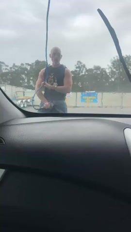 A bald man wearing a black tank top and blue shorts is holding a blue hose and a sprayer. He is standing in front of a car, and he appears to be spraying the car with water. The video is taken from inside the car, looking out through the windshield. The sky is cloudy, and there are trees in the background. There are also some trailers and a building in the background. The man is smiling and appears to be having fun.