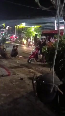 At night, a group of people are gathered on a dirt road, some sitting on mats, others standing. A red scooter is parked near the center of the frame, with a person sitting on it. There are buildings with lights in the background, suggesting a street or marketplace. Trees and foliage are visible, with some strings of lights hanging from branches. Several individuals appear to be interacting, though the specific nature of their actions is unclear due to the low light conditions and video quality.