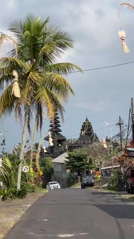 A vertical shot shows a road leading towards a dark, multi-tiered temple structure. Palm trees frame the left side of the shot, with long, decorative straw streamers hanging from curved poles on either side of the road. A white SUV is driving away from the camera on the road, and a dark SUV is parked to its right. The sky is cloudy.