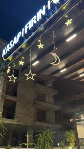 The video shows the exterior of a building at night. An illuminated sign that reads "KASAP I FIRIN I" is displayed prominently above the entrance, with decorative lights in the shape of stars and crescent moons hanging below. Behind the entrance, a partially constructed building with brick walls and empty window frames is visible. The sky is dark, and some greenery is visible near the base of the building, suggesting an outdoor setting.