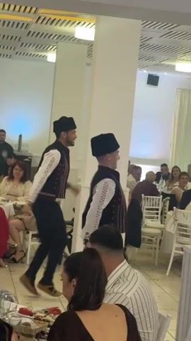 The video shows two men dancing in what appears to be a restaurant. Both men are dressed in traditional clothing including black vests and hats, and embroidered white shirts. One of the dancers kicks his leg up high. Several people are seated at tables surrounding the dancers, and many are watching them. The room has high ceilings, white columns, and several hanging lights. A woman with brown hair sits in the foreground facing away from the camera.