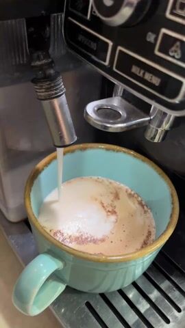 The video shows a light blue mug with a brown rim under the nozzle of a black and silver espresso machine. Milk is being dispensed into the mug. The mug already contains a coffee-colored liquid topped with foam. The espresso machine has buttons labeled "Cappuccino" and "Milk Menu." The mug sits on a black grate. The camera is positioned relatively close to the mug and espresso machine, providing a focused view.