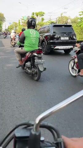 The media shows a street scene in Larangan, East Java, during daylight, likely late afternoon due to the warm lighting and long shadows. The weather appears clear and sunny.

The main subjects are individuals riding motorcycles on a paved road. One person in the foreground, on the left, is wearing a bright green jacket with the "Gojeck" logo and a black helmet, riding a white and red scooter. Another individual, in the center, also wears a green jacket with the Gojeck logo and a patterned helmet, riding a black and white motorcycle.

Several vehicles are present on the road. Behind the Gojeck riders, a black SUV is visible, followed by another black SUV. Other vehicles are present in the background, suggesting moderate traffic. The environment includes well-maintained roads, lush green trees and bushes lining the street, and streetlights. A traffic light is visible in the distance on the left.

The activity is daytime road travel, with the Gojeck riders seemingly engaged in delivery or transportation services. The overall atmosphere is that of a typical urban or suburban street scene.

Visible text includes the "Gojeck" logo on the green jackets and a license plate on the scooter in the foreground, which reads "H 6308 BDM".
