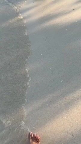 The video captures a close-up, top-down view of a person's feet walking on a wet sandy beach. The sand is light gray and appears damp, with some areas covered by shallow, clear water. Small waves gently wash over the sand, creating ripples and a foamy texture. Sunlight casts diagonal shadows across the scene, suggesting it might be late afternoon or early morning. The camera follows the person's feet as they move forward, with the water occasionally splashing around their ankles. The overall impression is one of a peaceful, tranquil walk along the shoreline.