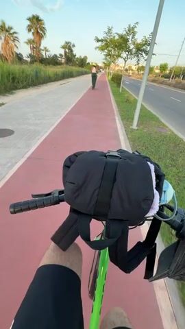 This first-person perspective video captures a cyclist riding on a dedicated red bike path alongside a road in Castelló de la Plana, Spain. The time of day appears to be late afternoon or early evening, indicated by the warm, golden light and the long shadows. The sky is a clear blue with a few wispy clouds.

The cyclist is equipped with a black handlebar bag, which appears to contain some items, including what looks like a blue face mask and a fluffy white object. The cyclist's legs are visible, wearing black shorts. The bike itself is bright green.

In the distance, another person is riding what appears to be an electric scooter or bike on the same path, moving away from the camera. Palm trees and other vegetation line the path and the roadside, suggesting a warm climate. A paved road with a single lane of traffic runs parallel to the bike path. The overall mood is peaceful and relaxed, suggesting an enjoyable ride in pleasant weather. There is no visible text in the video.