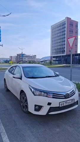 The video opens with a close-up of a white Toyota Corolla parked on a paved surface, likely a street or parking lot. The car is facing the camera, with its front bumper and grille clearly visible. The license plate reads "B619HA 89". The scene is outdoors, with a clear blue sky and scattered clouds suggesting it's daytime.

As the camera pans to the right, a modern, tall building with a pinkish-brown facade and numerous windows comes into view. A triangular yield sign is mounted on a pole in front of this building. Further in the background, another similar tall building is visible, along with a lower, grey building with a distinctive green, sloped roof feature. The overall setting appears to be an urban or developing area, with modern architecture and well-maintained roads.

The camera continues to pan, revealing more of the street and sidewalk. There are bollards with red and white markings on the sidewalk, suggesting a pedestrian area or parking demarcation. The environment looks clean and orderly, and the lighting indicates it's likely late afternoon or early evening, given the soft sunlight. No people are clearly visible in the frame, though a faint silhouette can be seen in the driver's seat of the car. The overall mood is calm and observational, with a focus on the urban landscape and the parked vehicle. The location cues, such as the building styles and the Russian license plate format, point to a modern city in Russia, specifically Novy Urengoy as indicated.