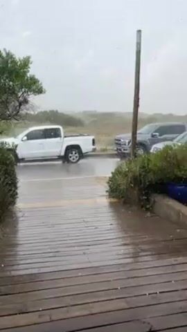 The video shows a heavy rainstorm hitting a beachside area with several parked cars. The camera is positioned on a wooden deck overlooking a paved road. A white pickup truck and a silver SUV are parked on the road, partially visible through the heavy rain. Behind them, another dark-colored vehicle is parked. The rain is so intense that it creates a dense white haze, obscuring the view of the beach and sand dunes in the background. The wooden deck is wet and glistening. The video focuses on the driving rain and the parked vehicles. The overall scene depicts a harsh weather event impacting a coastal area.