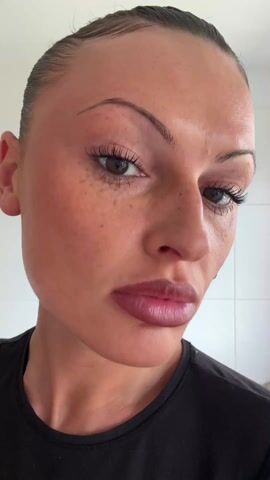 A fair-skinned woman with freckles and dark hair pulled back is applying black mascara to her upper eyelashes. Her face is close to the camera, and she is looking down slightly as she carefully coats her lashes. She is wearing a black shirt. The background appears to be a white tiled wall, possibly in a bathroom.