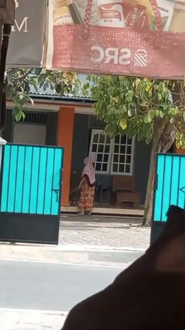 A woman wearing a pink headscarf and a floral-patterned dress is sweeping the paved area in front of a house. She is holding a broom and appears to be working. The house has an orange and gray facade, a white-paned window, and a porch. Two blue gates, one open and one partially open, are visible in the foreground. A tree with green foliage is situated to the right of the house. A banner with red and white text, partially obscured, is visible at the top of the frame. The scene is outdoors, under daylight.