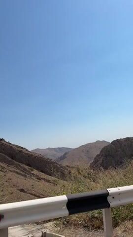 The video shows a view from a moving vehicle traveling along a mountainous road. The foreground features a white and black guardrail. Beyond the guardrail, a dry, grassy hillside slopes upward. In the distance, more rugged, arid mountains are visible under a clear blue sky. The scene suggests a sunny day in a mountainous, possibly desert or steppe environment.