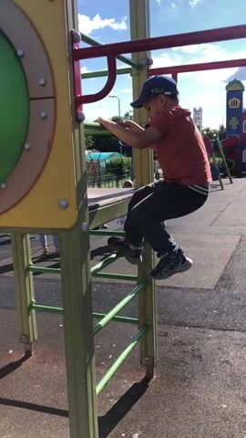 A young boy wearing a red t-shirt, gray pants, and a blue baseball cap is climbing a playground structure. He is using a ladder-like structure with green rungs to ascend. He has his hands on the rungs and his feet on the rungs as he climbs. The playground structure is yellow and green. There are trees and a blue sky in the background.