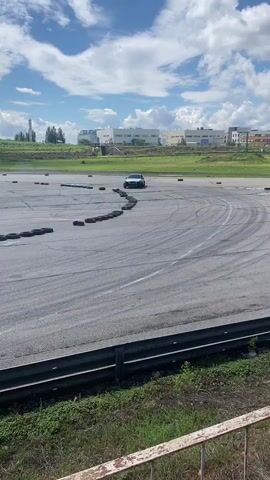 This media file captures a drifting event on a sunny day in Usol'e, Russia. The primary subject is a dark-colored car, possibly a sedan, engaged in a drift. The car is kicking up a significant amount of tire smoke as it maneuvers around a track marked by a series of tires.

The scene is an outdoor asphalt track with a clear blue sky dotted with white clouds. In the background, there are several industrial-looking buildings and some distant trees. A power line and utility pole are also visible. The track itself has white lines painted on the asphalt and a barrier made of what appears to be metal fencing in the foreground. Lush green grass and weeds grow along the fence.

The time of day appears to be daytime, indicated by the bright sunlight and shadows. The weather seems pleasant, with a bright sky and scattered clouds. The overall atmosphere suggests an energetic and exciting event, likely a race or a demonstration of drifting skills.

Visible text on the car includes what appears to be "MAKITA" and "DRIFT" in stylized fonts, suggesting a sponsorship or branding related to the event. The car also has what looks like a racing number or logo on its side. There is also a small figure of a person standing in the background on the track, observing the action.