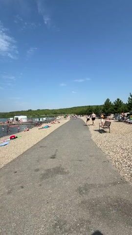 The video shows a wide shot of a beach and a body of water. A paved walkway runs along the edge of the beach, with a gravelly shoreline sloping down to the water. Numerous people are visible on the beach, some sunbathing, others walking. In the water, many people are swimming and playing. In the distance, there are buildings and trees, and the sky is clear blue with a few white clouds. There are also large, golden letters spelling "BOSS" visible in the water in the mid-ground. The overall scene depicts a lively and sunny day at a beach resort.
