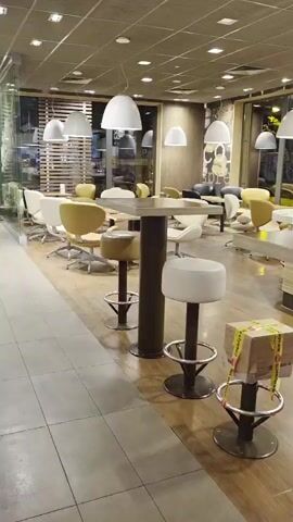 The video shows the interior of a McDonald's McCafe area. Several high-top tables with bar stools are visible. One of the bar stools in the foreground has a cardboard box on it, secured with yellow and black caution tape. The McCafe counter is visible in the background, with a display case containing food items and a cash register. The lighting is dim, and the overall atmosphere appears to be quiet and empty, suggesting the establishment may be closed or in its off-peak hours.