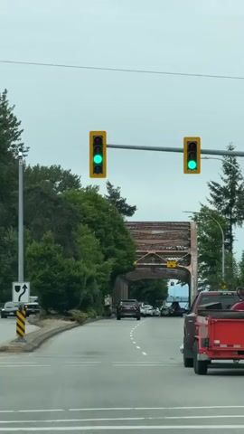 The video shows a street scene with a car's perspective from the driver's seat. Two traffic lights are visible, both displaying a green light. A red pickup truck with a flatbed trailer is in the right lane. Ahead, a bridge with a rusty metal structure spans across the road. Several cars are visible on the road, some driving under the bridge. Lush green trees line the left side of the road, and a few tall pine trees are on the right. The sky is overcast. There is a yellow sign with black text visible on the bridge structure, indicating a clearance height of 4.5 meters.