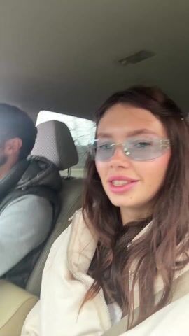 A young woman with long brown hair, wearing reflective silver sunglasses and a cream jacket, sits in the passenger seat of a car. She looks at the camera and speaks. The male driver, who is wearing glasses, a gray hooded sweatshirt, and a dark gray vest, sings and gestures emphatically. The car is in motion, as suggested by the blurring background. The car's interior is visible.