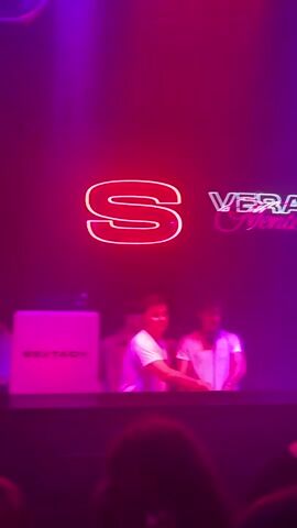 The video shows three young men who appear to be DJs at a nightclub or performance venue. They are behind a mixing console, and neon signs above them spell out "S" and "Vera Niente." There's also a box in the lower left corner that says "SEXTACY." Two of the DJs are wearing white t-shirts, and the one on the right has headphones around his neck. The lighting in the venue is dark, and the dominant colors are red and purple. A fourth person is standing in the foreground facing away from the camera, obscuring the full view.