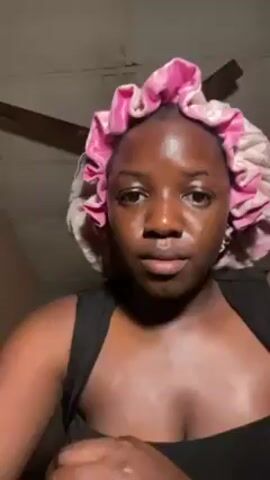 A young, dark-skinned woman with visible facial oil is applying lip liner. She's wearing a pink-and-white floral bonnet and a black tank top. She holds a gold lip liner pencil and carefully traces the outline of her lips. The background includes a ceiling fan and a neutral-colored wall.