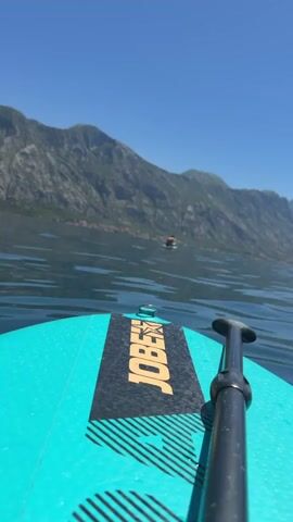 The video shows a point-of-view shot from a stand-up paddleboard (SUP) on a body of water. The board is teal and has a black stripe with the word "JOBE" in yellow on it. A paddle rests on the board. In the distance, there are mountains and a clear blue sky. A person is visible in the water further away from the camera.  The water is calm with slight ripples.