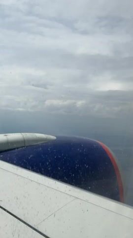 The video shows a view from an airplane window. The left side of the frame is dominated by a white airplane wing with some smudges or droplets on it. To the right of the wing is the engine nacelle, which is primarily dark blue with small white speckles and a red stripe at its edge. The background consists of a cloudy sky with varying degrees of light and dark, suggesting a partly cloudy day. The horizon is visible in the distance, showing a landmass with some faint details, possibly fields or a settlement, but it is obscured by atmospheric haze. The camera appears to be relatively still, with no significant panning or tilting, indicating a static shot from inside the aircraft. The overall impression is a snapshot of a flight in progress, emphasizing the mechanical components of the aircraft and the atmospheric conditions outside.