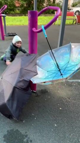 The media file shows a young child kneeling on a playground. The child is wearing a grey jacket, blue pants, green rubber boots, and a patterned cap. In front of the child are a toy tractor and a small green toy truck. Two open umbrellas are propped up near a piece of playground equipment. One umbrella is black and the other is a light blue color with a design on it. The playground equipment is pink and purple. The ground is asphalt. In the background, there are trees, buildings, and parked cars. The lighting suggests it is daytime. The scene appears to be taking place outdoors in a residential area, possibly a courtyard or a public park. The child's focus on the toys suggests they are engaged in play. The presence of umbrellas might indicate recent rain or a chance of rain, although no precipitation is visible in the footage.