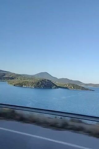 The video is a shaky, horizontal shot of a coastal landscape seen from a moving vehicle. The left side of the frame shows a body of blue water with islands and hills in the background. The right side of the frame is a blur of green foliage and brown rocks. A gray road with a white line is visible in the foreground, indicating the perspective of someone traveling in a car. The sky is clear and blue. The footage appears to be taken during daylight.