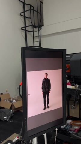 The video shows a man getting his photo taken in a studio setting. The first shot shows the monitor displaying a test shot of a man in a black jacket and black pants against a white background. The next shot shows two people working at desks, one with a laptop and the other with a phone. The next shot shows a stool, a stand, and a tan cylinder prop in a studio space. The final shot shows a man standing in the studio, wearing a black and grey jacket, black pants, and red-accented black shoes. He is looking to his left. The studio has a white backdrop, a door, and a fan in the background.