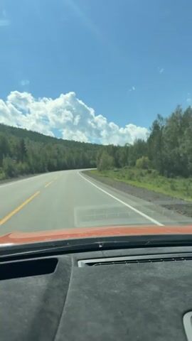 The video begins with a view from inside a vehicle looking out through the windshield. The dashboard is visible in the foreground. The road ahead is a paved two-lane highway with a yellow center line and a white edge line. The road curves to the right, surrounded by a dense forest of green trees. In the distance, there are rolling hills also covered in trees. The sky is a clear blue with a few fluffy white clouds. Sunlight streams from the left, creating a lens flare effect on the windshield. The car appears to be in motion, traveling along the highway. A red and white circular road sign is partially visible on the right side of the road. The overall environment suggests a scenic drive through a wooded, possibly mountainous, region on a sunny day.